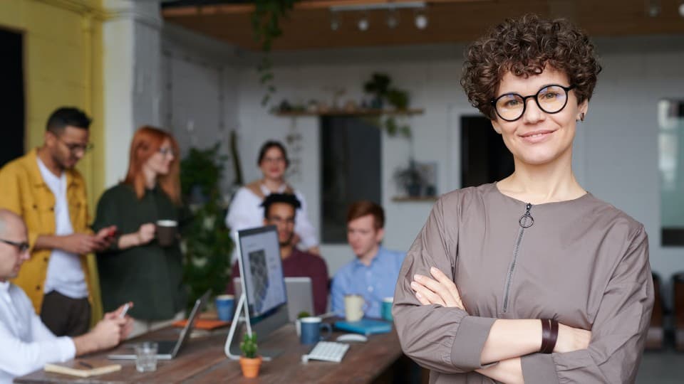 Nainen seisoo kädet ristissä katsoen kameraan ja ihmisiä istuu pöydän ääressä